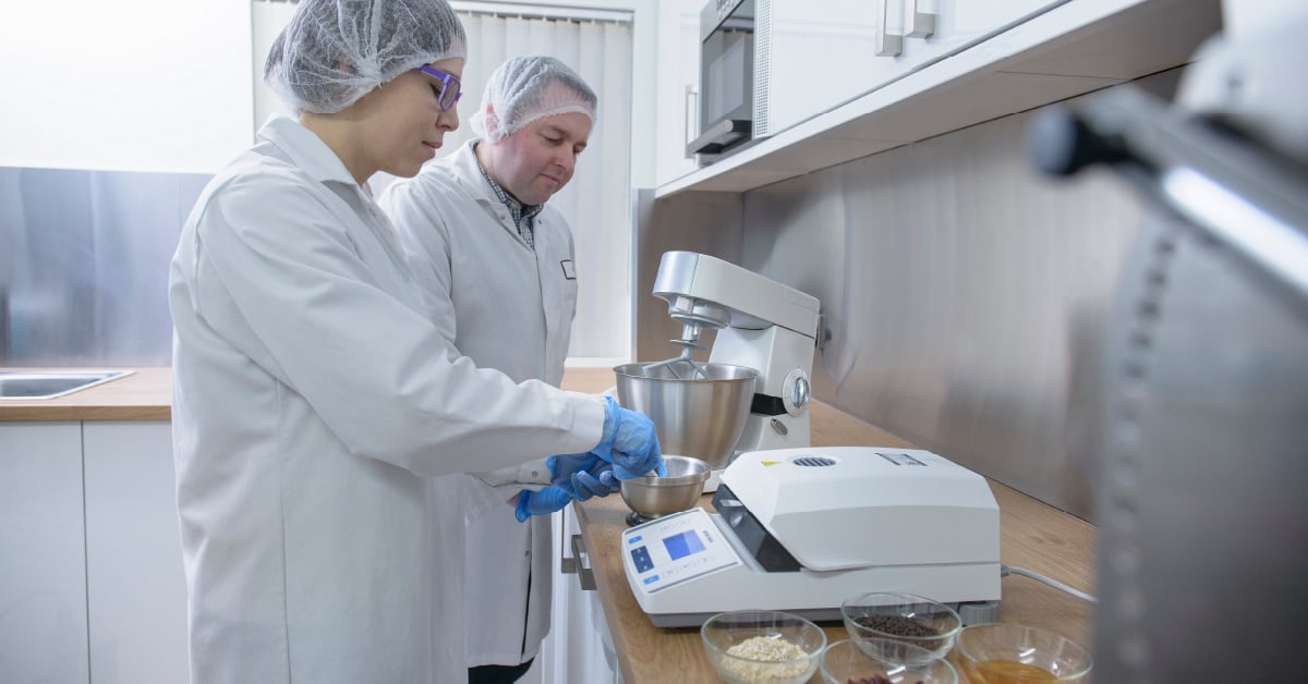 Two food manufacturers in smocks work with ingredients in a pilot test kitchen